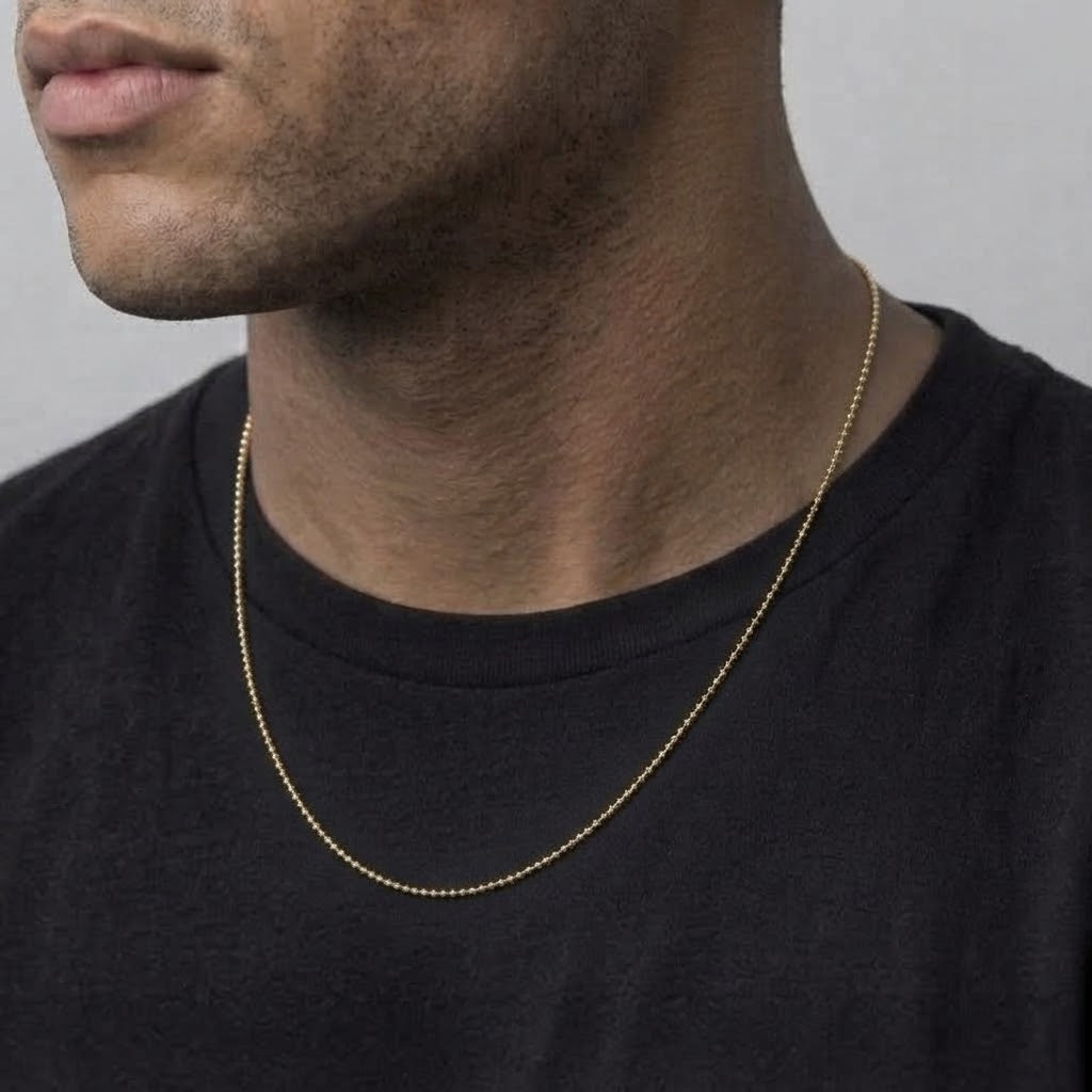 A close-up of a man wearing a delicate gold chain necklace over a black t-shirt, showing the necklace from the front against a neutral gray background.