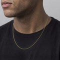 A close-up of a man wearing a delicate gold chain necklace over a black t-shirt, showing the necklace from the front against a neutral gray background.