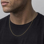 A close-up of a man wearing a delicate gold chain necklace over a black t-shirt, showing the necklace from the front against a neutral gray background.