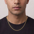 A close-up of a man wearing a delicate gold chain necklace over a black t-shirt, showing the necklace from the chest up against a light gray background.