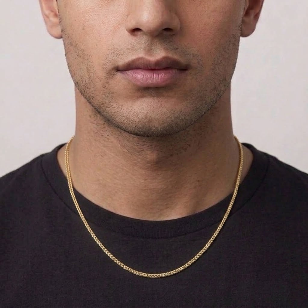 A close-up of a man wearing a delicate gold chain necklace over a black t-shirt, showing the necklace from the chest up against a light gray background.