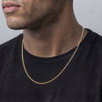A close-up of a man wearing a delicate gold chain necklace over a black t-shirt, showing the necklace draped around his neck.