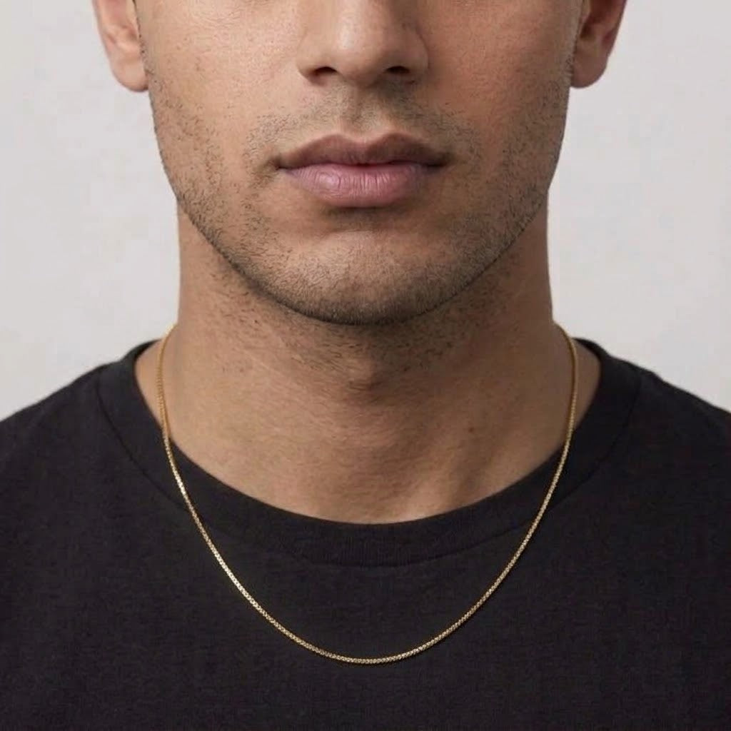 A close-up of a man wearing a delicate gold chain necklace over a black t-shirt, showing the necklace draped around his neck.