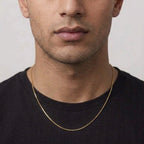 A close-up of a man wearing a delicate gold chain necklace over a black t-shirt, showing the necklace draped around his neck.