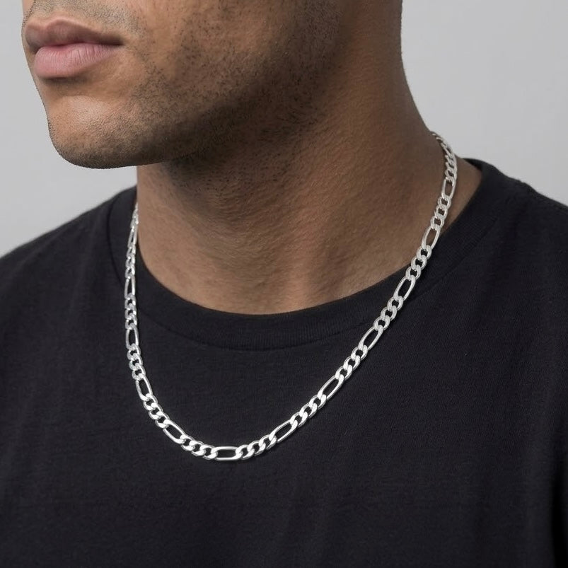 A close-up of a man wearing a silver Figaro chain necklace over a black t-shirt, showing the necklace against his neck.