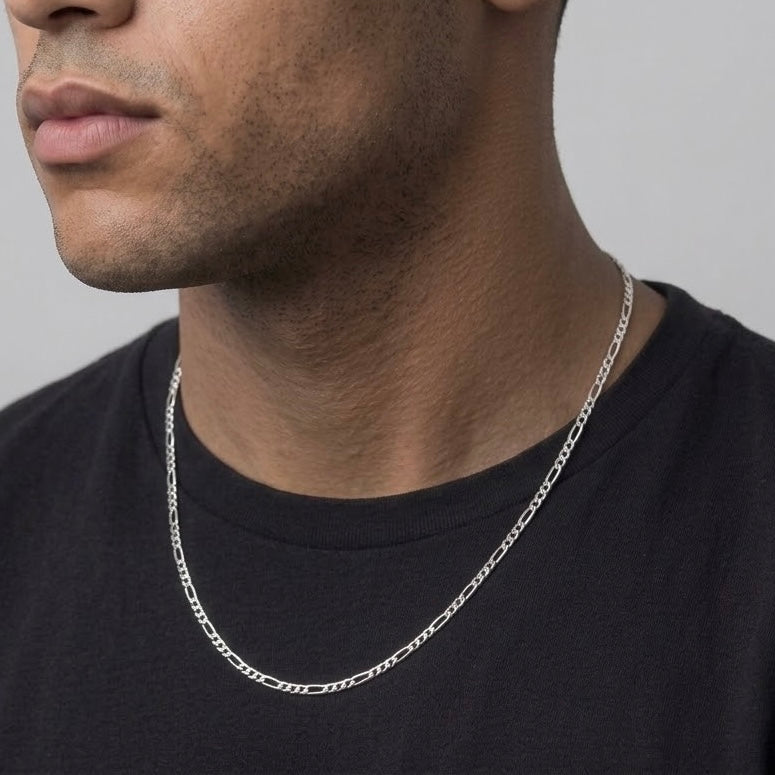 A close-up of a man wearing a silver Figaro chain necklace over a black t-shirt, showing the necklace against his neck.