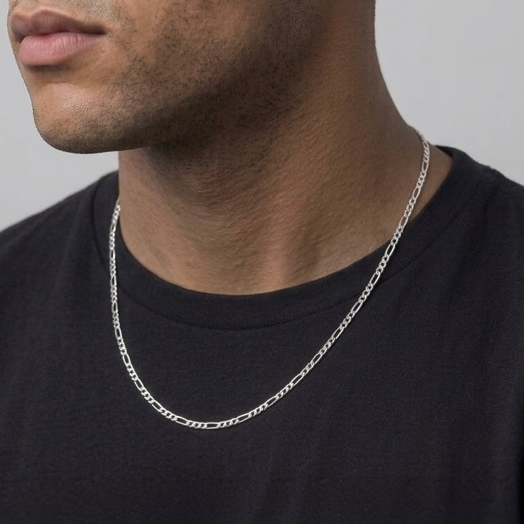 A close-up of a man's neck and lower face wearing a thin silver Figaro chain necklace over a black t-shirt, against a plain light gray background.